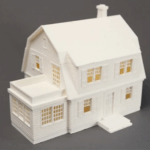 Weißes Modell eines zweistöckigen Hauses mit Satteldach und Schornstein, verglastem Anbau mit vielen Sprossenfenstern, horizontaler Fassadenvertäfelung und kleinem Vordach vor schlichtem grauem Hintergrund.