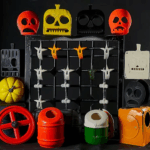 Verschiedene bunte Kunststoff-Spielzeugteile in Halloween-ähnlichen Formen – mehrere Totenkopfmasken in Rot, Gelb, Schwarz und Orange, Kürbis- und Rad-Elemente sowie eine schwarze Rastertafel mit kleinen weißen und orangen Totenkopffiguren – arrangiert vor schwarzem Hintergrund.