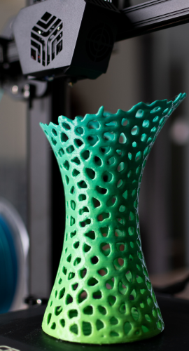 Grün-türkisfarbene, gitterartige Vase mit löchriger, geschwungener Form steht auf dem Bauplateau eines 3D-Druckers, dessen Extruder im Hintergrund sichtbar ist.