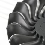 Nahaufnahme eines schwarzen, wellenförmigen Kunststoff-Lüfterrads mit mehreren geschwungenen Flügeln und sichtbarer feiner Schichtstruktur auf hellem Hintergrund.