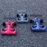 Drei spielzeug-Formelrennwagen in Rot, Grau und Blau mit Heckflügeln und breiten Reifen nebeneinander auf rauem Asphalt platziert.
