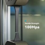 Zugprüfmaschine dehnt einen schwarzen, schichtartig wirkenden Kunststoff-Probestab bis zum Bruch; rechts eingeblendet der Text ‚Tensile Strength 100Mpa‘.