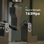 Dreipunkt-Biegeprüfung einer schwarzen Probenleiste in einer Prüfmaschine, bei der ein mittiges Eindrückwerkzeug die Probe auf zwei Auflagestützen drückt; eingeblendeter Text zeigt Flexural Strength 163 MPa.