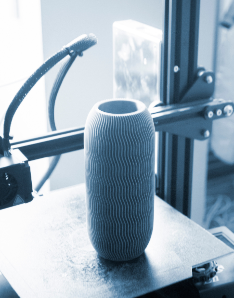 Zylindrische 3D-gedruckte Vase mit gleichmäßigen wellenförmigen Längsrillen steht auf der Bauplatte eines 3D-Druckers, im Hintergrund sind der Metallrahmen und eine flexible Leuchte zu sehen.