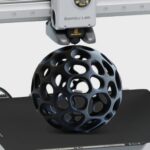 Bambu Lab 3D-Druckkopf über dem Druckbett, der eine schwarze, hohle Kugel mit organisch geformten, ovoiden Öffnungen (Voronoi-ähnliches Gitter) druckt.