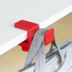 Roter Kunststoffhaken, an der weißen Tischkante eingeklemmt, trägt den grauen Träger einer Stofftasche vor hellem Holzboden.
