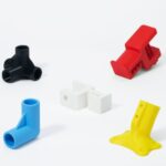 Sechs farbige Kunststoff-/3D‑gedruckte Verbindungsteile—schwarzer dreifacher Rohrverbinder, rotes Klemmelement, weißes zweigeteiltes Steckstück, blaues 90°-Winkelrohr mit Stütze und gelbe geschraubte Winkelplatte—auf weißem Hintergrund angeordnet.