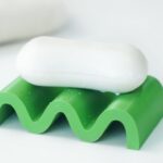 Weißes Stück Seife liegt auf einem grünen, wellenförmigen Seifenhalter mit sichtbaren Wassertropfen, vor einem unscharfen weißen Handtuch.