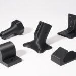 Sechs schwarze, mattierte Metall-/Kunststoff-Befestigungsteile unterschiedlicher Formen (u. a. L-Winkel mit mehreren Schraublöchern, zylindrische Rohrschelle, abgeschrägter Halter und konisches Muffenstück) liegen auf weißem Hintergrund.