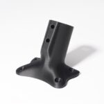 Schwarze, schräge Metallflansch-Halterung mit strukturierter, matter Oberfläche, vier Bodenbefestigungsbohrungen und zwei Schraubenlöchern in der geneigten Rohraufnahme auf weißem Hintergrund und leichtem Schatten.