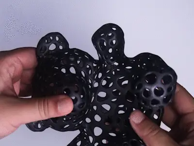Zwei Hände halten vor hellem Hintergrund ein schwarzes, organisch geformtes 3D‑Gitterobjekt mit zahlreichen runden Öffnungen.