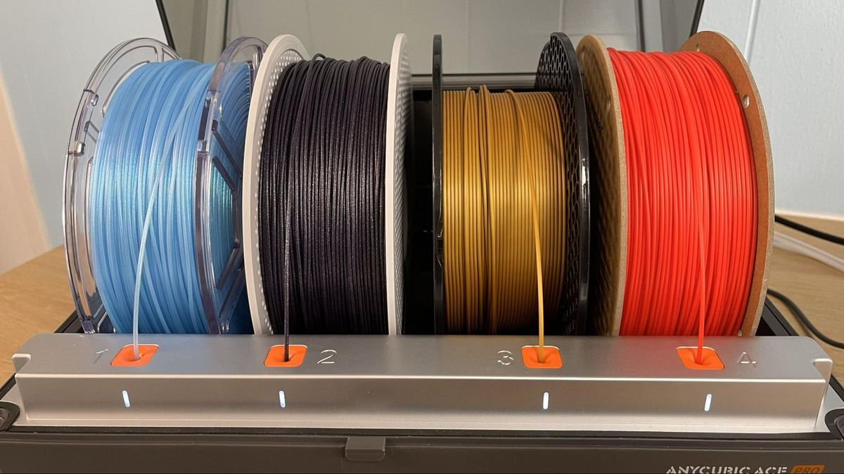 Vier Spulen 3D‑Druckfilaments in Hellblau, Schwarz, Gold und Rot nebeneinander auf einem silbernen Spulenhalter mit vier orangefarbenen Einzugslöchern (nummeriert 1–4) und weißen Status‑LEDs, auf einem Tisch vor einem Drucker.