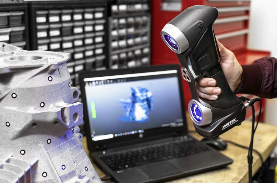 Handgehaltener 3D‑Scanner mit blauen Lichtquellen wird auf ein mit runden Markern versehenes gegossenes Metallgehäuse gerichtet, während ein Laptop im Hintergrund das digitale Scanmodell anzeigt und sich Schubladen einer Werkstatt befinden