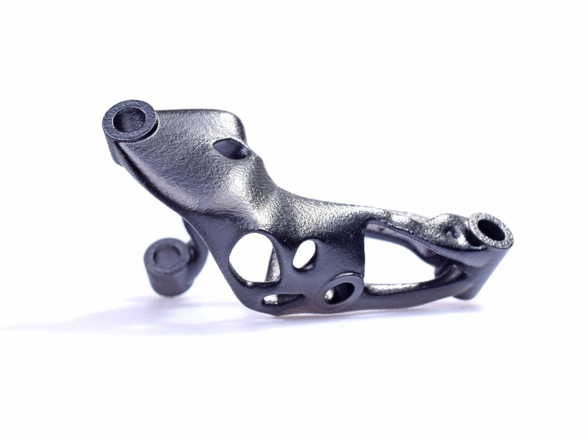 Schwarz-graues, organisch geformtes Metallbauteil mit mattem Finish, mehreren runden Durchbrüchen und zylindrischen Aufnahmen an den Enden auf weißem Hintergrund.